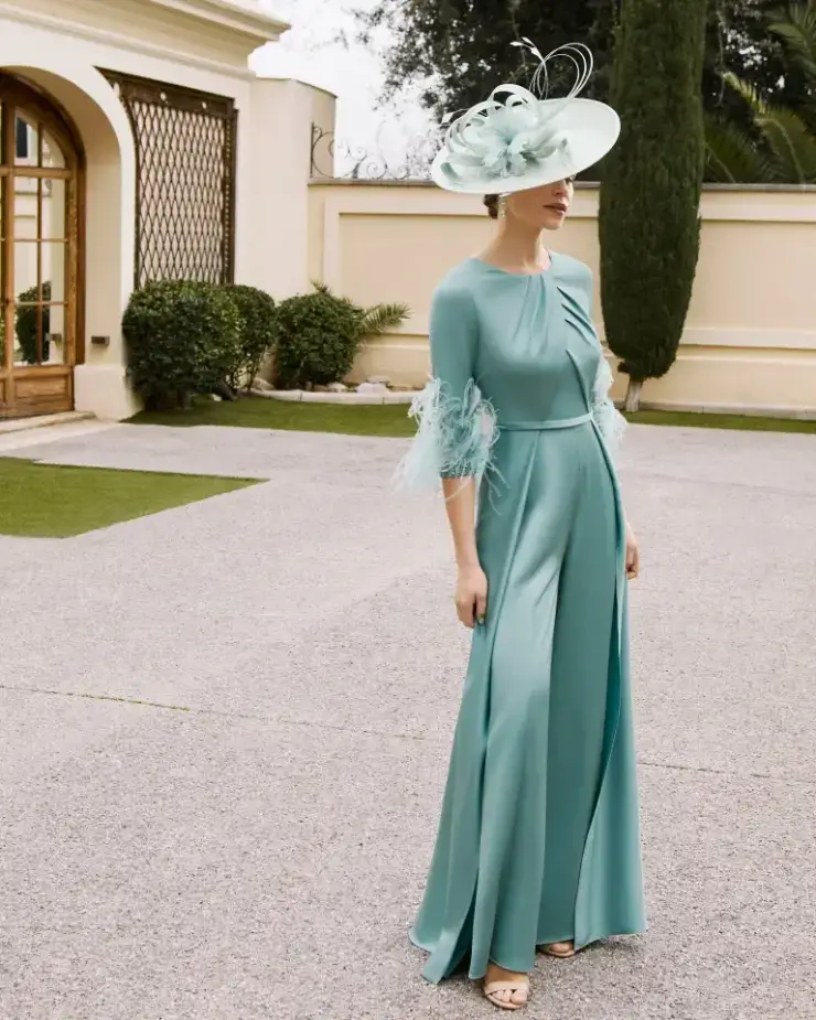 A woman wearing a teal dress and a large decorative hat, standing outdoors on a paved area with greenery in the background.