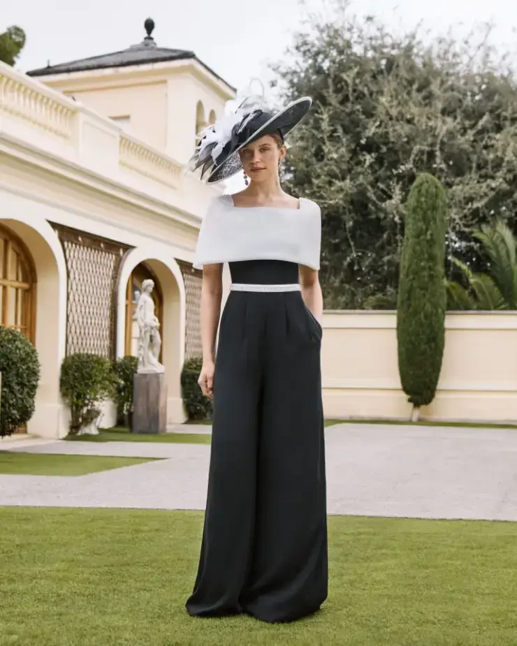 A woman in a black and white formal outfit stands outside a building, wearing a large, decorative hat.