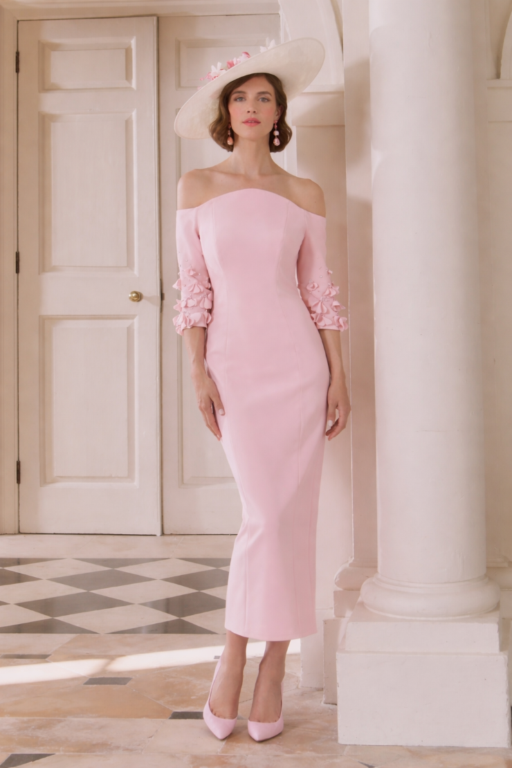 A woman wearing a pink off-the-shoulder dress with three-quarter sleeves and floral embroidery, complemented by a wide-brimmed white hat and pink heels, standing elegantly in a spacious indoor setting.