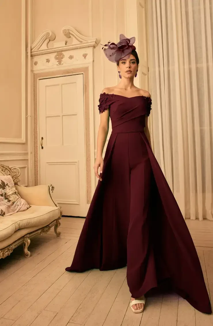 A woman in a burgundy off-shoulder gown with a flowing train, complemented by a large headpiece, walking gracefully in a beautifully decorated room.