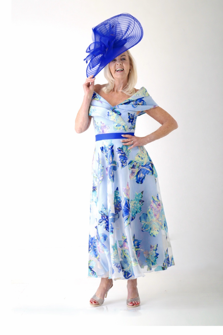 A woman wearing a floral blue dress and a large blue hat, posing confidently.