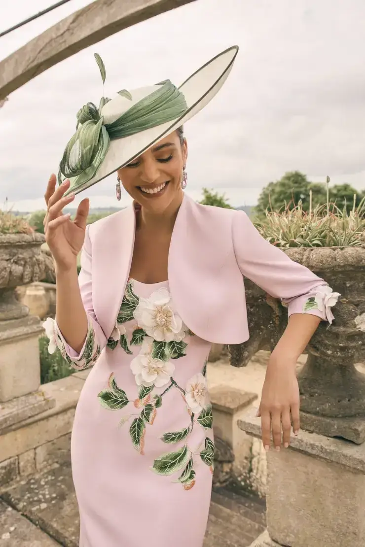 A woman wearing a pink dress adorned with floral embroidery and a matching cropped jacket, accompanied by an oversized decorative hat. She is smiling while posing outdoors near an ornate stone structure.