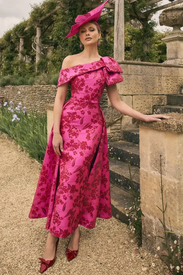 A woman wearing a pink floral dress and a matching hat stands elegantly by a stone wall and staircase, surrounded by greenery and flowers.