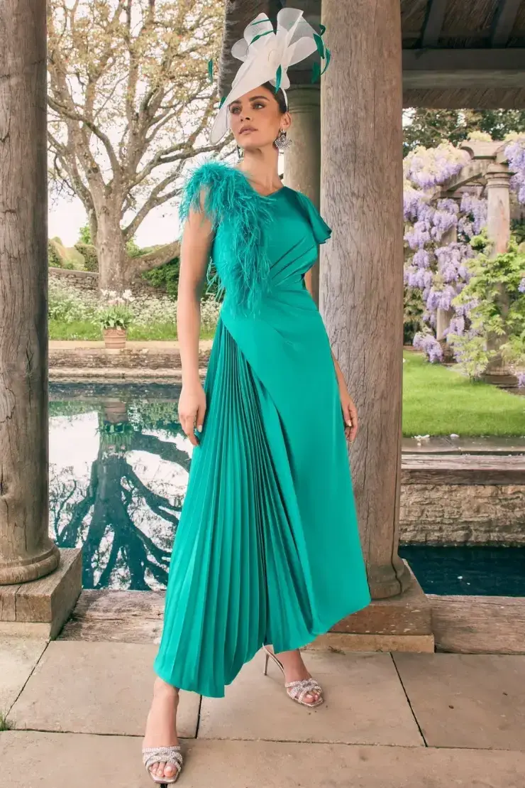 A model wearing an elegant turquoise dress with feathered detailing and a flowing pleated design, standing outdoors near a water feature surrounded by greenery and blooming flowers. The model is accessorized with a stylish hat.