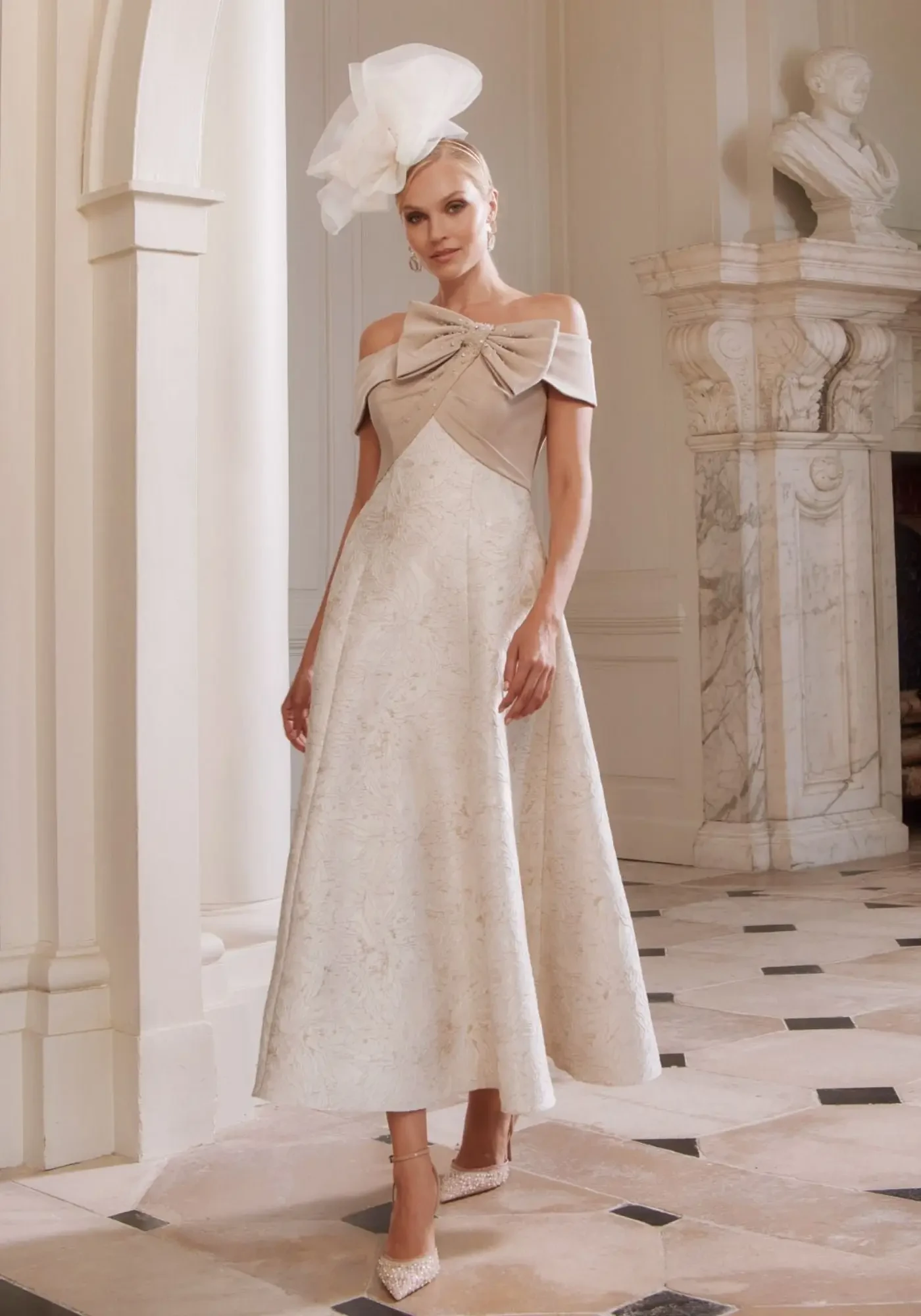 A woman is wearing an elegant off-the-shoulder dress with a large bow detail at the neckline. The dress is cream-colored with subtle patterns and falls to mid-calf. She has styled her hair elegantly and is accessorized with a large decorative headpiece, standing in a grand, marble-floored room.
