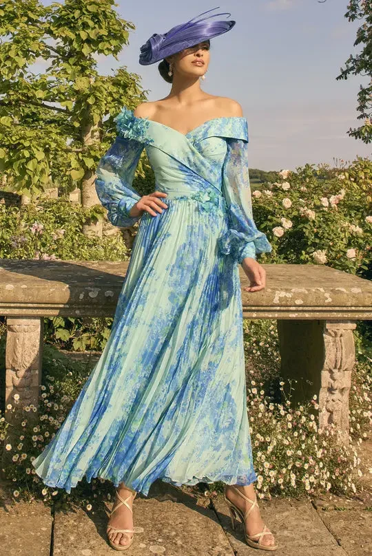 A model wearing a flowing blue and green dress with an off-the-shoulder design and sheer sleeves, paired with a large purple hat, stands in a garden filled with flowers.