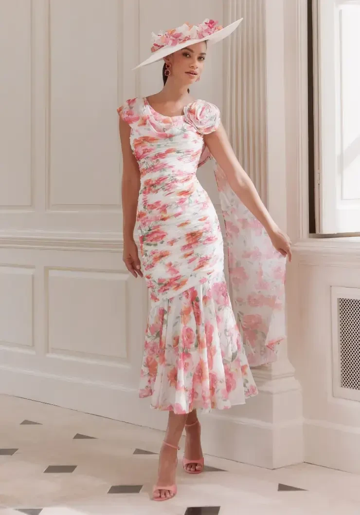 A woman in a floral dress with ruffles and a wide-brimmed hat poses elegantly in an indoor setting.