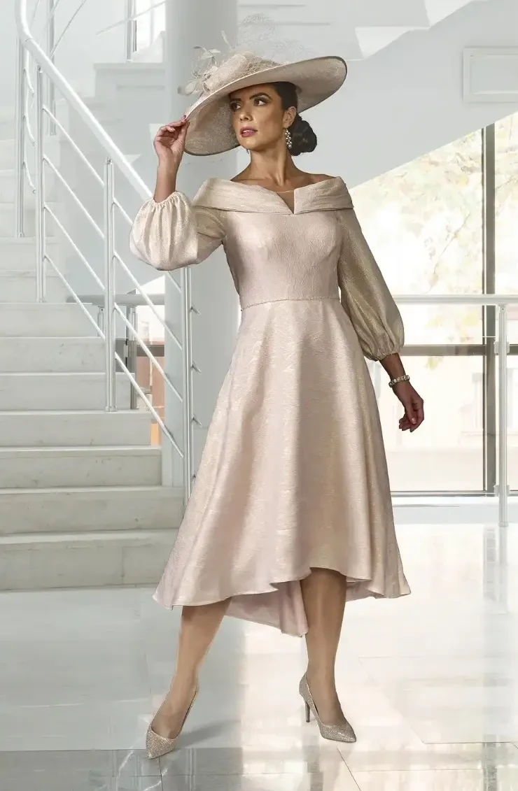 A woman in a light-colored elegant dress with three-quarter sleeves and a high-low hem, wearing a wide-brimmed hat and heels, posing on a staircase.