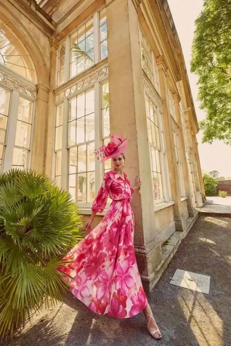 A woman in a pink floral dress poses elegantly outside a building with large windows, surrounded by greenery.